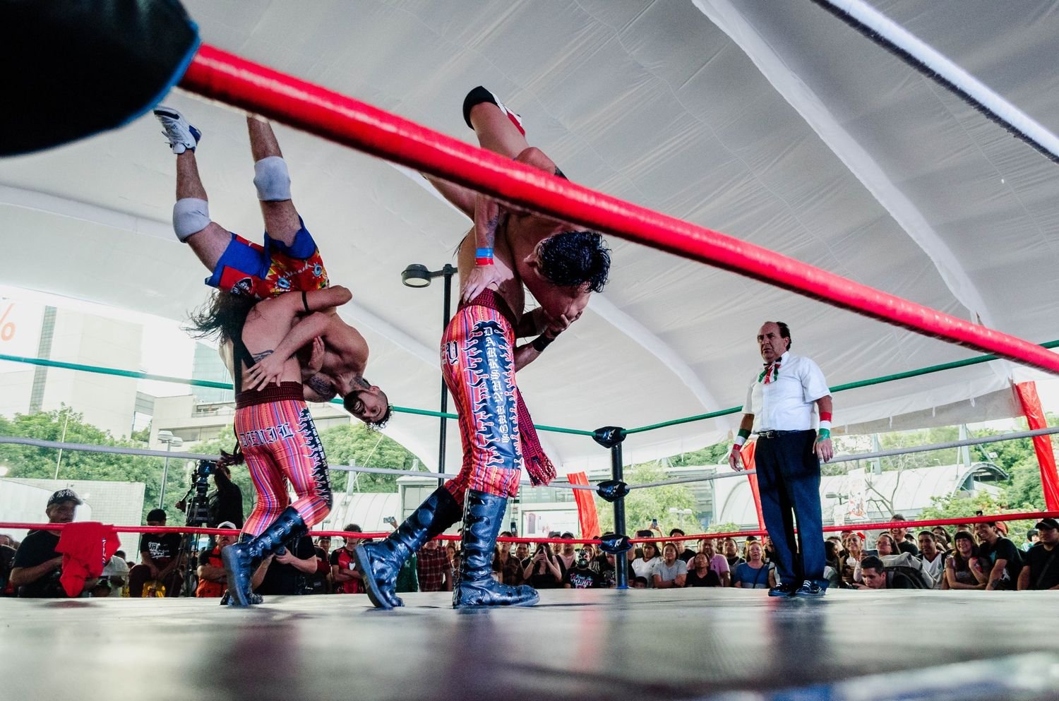 Dos luchadores ejecutando una maniobra en el aire bajo una carpa mientras el réferi observa